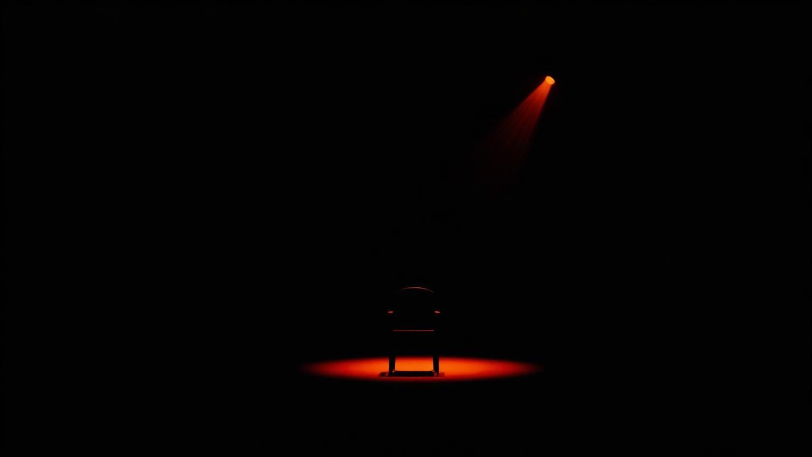 An empty chair under a single ember spotlight on a dark stage